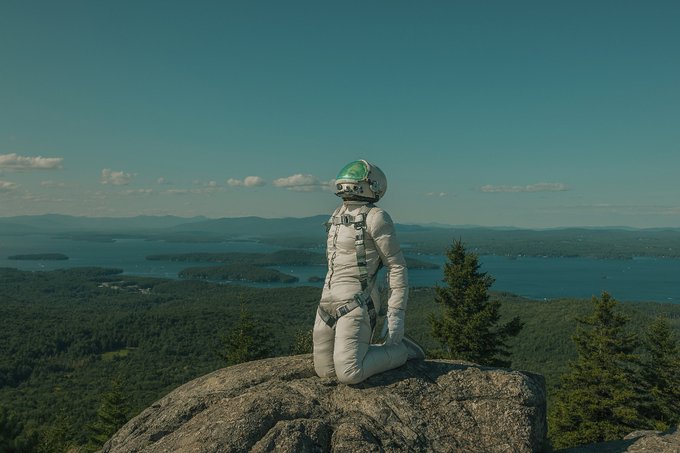 An astronaut in a white spacesuit with a green-visored helmet kneels on a rocky mountain peak, overlooking a vast landscape of forested hills and a large lake under a blue sky.