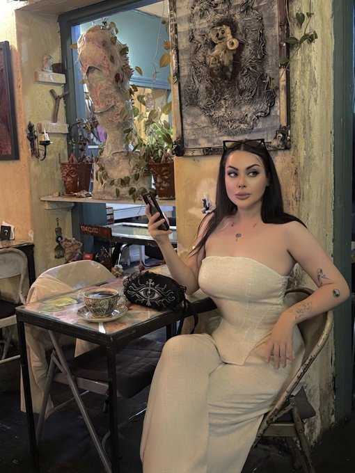 A woman with long dark hair wearing a strapless cream-colored dress sits at a small bistro table in a vintage-styled café, holding a smartphone while surrounded by ornate wall art and decorative objects.