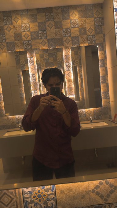 A mirror selfie of a man in a maroon shirt holding a smartphone in a bathroom with patterned Mediterranean-style tile walls.