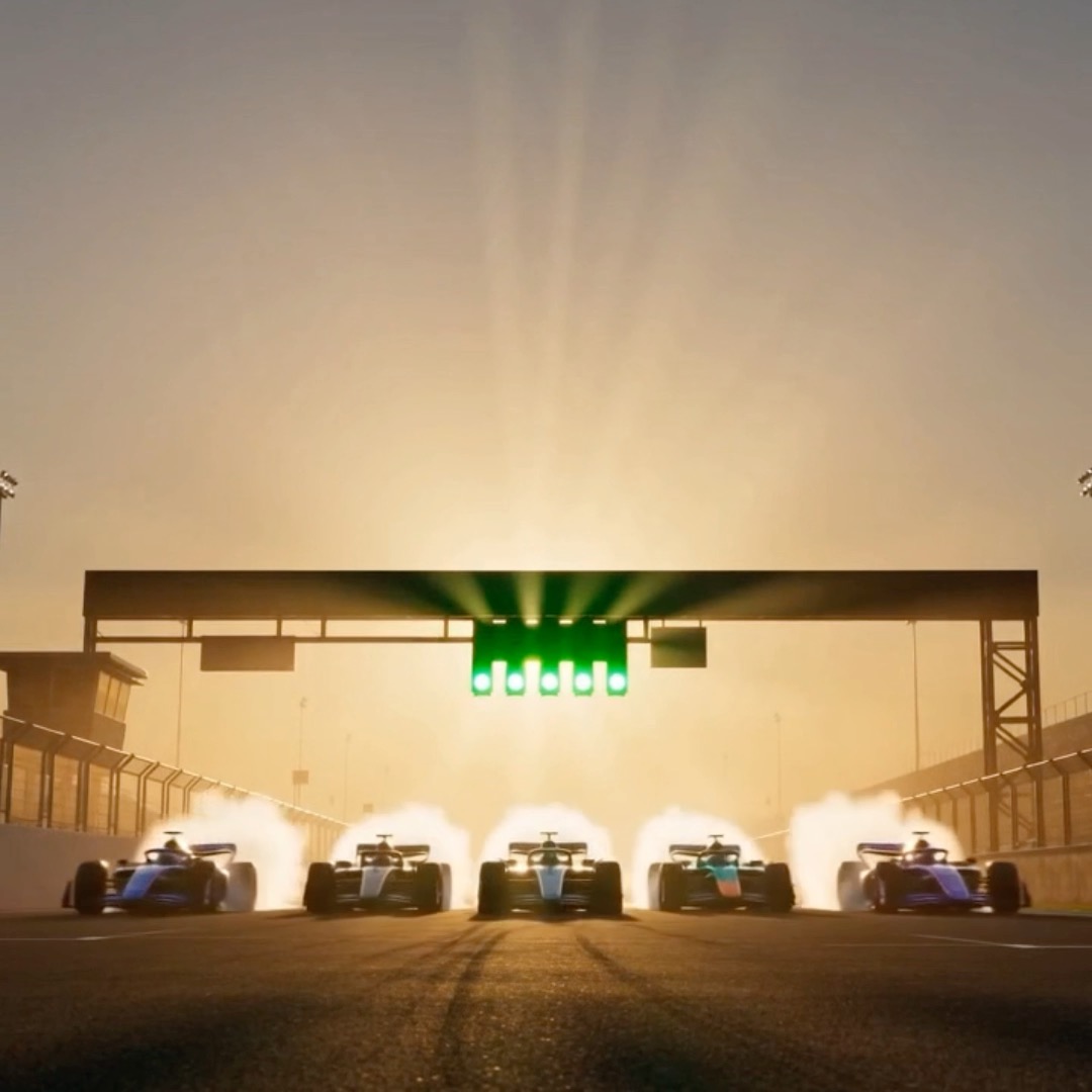 Five Formula race cars line up at a starting grid with a green light illuminated above, bathed in a dramatic golden light as exhaust or smoke billows from the vehicles.