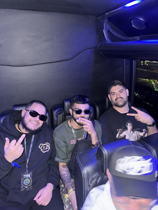 Three men posing inside what appears to be a vehicle or lounge, with one man in the foreground wearing sunglasses and making a hand gesture, and another wearing a 'TYPE' shirt.