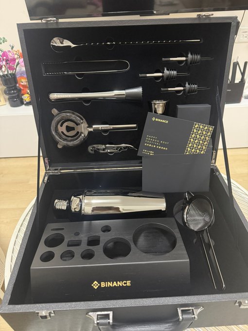 The image features a black briefcase filled with various cocktail shakers, glasses, and other accessories. The briefcase appears to be open, revealing its contents. A note attached to the briefcase reads "我也收到来自币安的VIP端午节礼物了", which suggests that someone has sent these cocktail shakers as a gift or surprise for another person, possibly a VIP.

The image also includes some text and objects related to the word "binance". There is a chair in the background, along with a potted plant nearby. The presence of these items further emphasizes the theme of the image - cocktails and mixology.