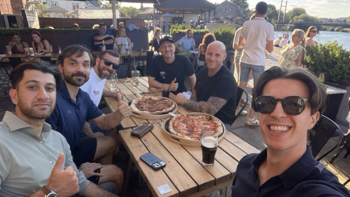 A group of five men pose for a selfie around a wooden table with two pizzas and drinks at a waterfront restaurant, one man flashing a thumbs-up.