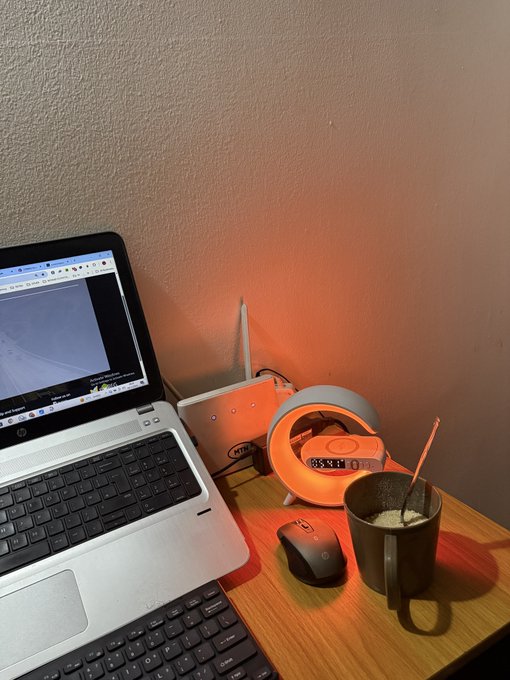 A laptop displaying charts sits on a wooden desk next to a glowing orange alarm clock showing "5:48," a wireless mouse, and a mug of coffee. The "Gm to all my pre-rich frens" caption fits perfectly as a morning greeting from a trader or investor getting an early start on market analysis, with the pre-dawn alarm and lit-up screen suggesting dedication to catching market moves.