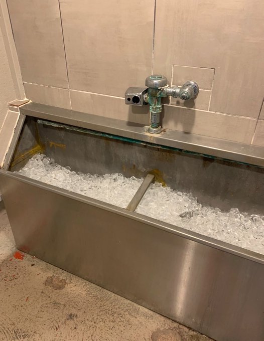 A stainless steel trough-style drinking fountain filled with crushed ice sits against a tiled wall, equipped with a metal spigot and push button.