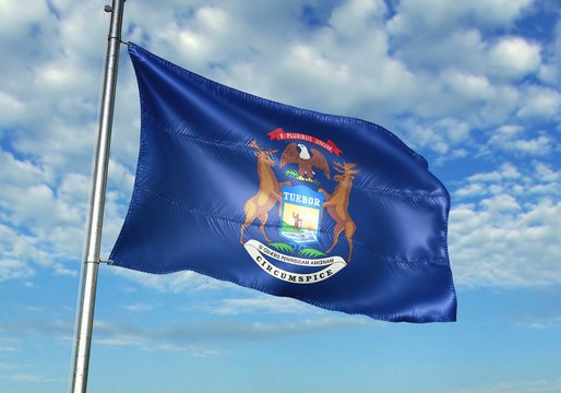 The flag of Michigan waving against a blue sky with clouds, featuring its coat of arms with elk and moose, and the Latin motto 'TUEBOR' and 'CIRCUMSPICE'.