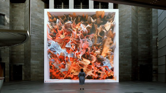 The image features an art installation with a large screen displaying a digital painting, which appears to be a complex piece of abstract art composed of various shapes and colors. A woman stands in front of this artwork, observing it closely. The surrounding area includes several doors and windows, suggesting that the space is likely part of a larger building or exhibition venue.