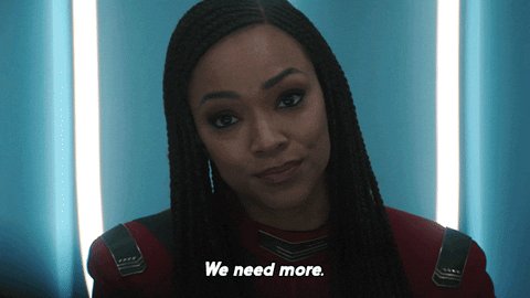 The image features a woman with long braids wearing a red shirt, standing against a blue background. Below her head, there's text that reads "We need more", which appears to be part of an advertisement or promotional material for a product or service. The focus on this message and the woman's presence in front of it suggests that she is being used as a visual representation of the advertised item or concept.