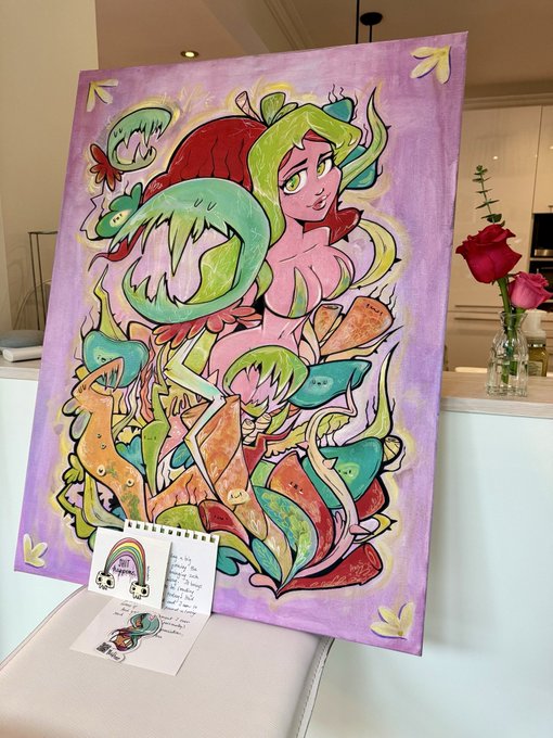 A colorful painting on a canvas featuring stylized green and red characters and abstract plant-like forms against a purple background, sitting on a counter next to a small handwritten note card.
