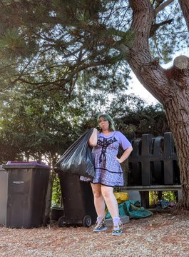A woman wearing a patterned dress stands between black garbage bins while holding a large trash bag outdoors near trees.
