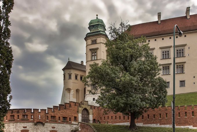 A historic Polish castle or palace featuring a prominent tower with a green copper roof, cream-colored walls, and a fortified brick base, set against a dramatic cloudy sky with a large tree in the foreground. The post has no direct connection to the image, which appears to be a scenic travel photo from Kraków rather than related to the Abstract blockchain or AI agents.