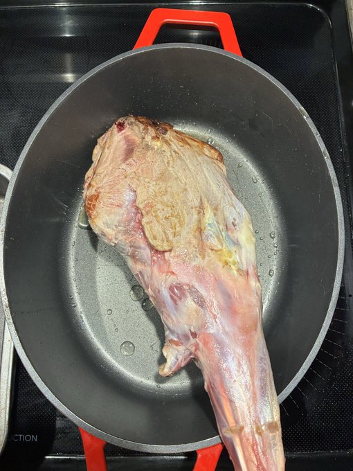 A large raw leg of meat, possibly lamb or goat, searing in a black cast iron pan with red handles on an induction stovetop.