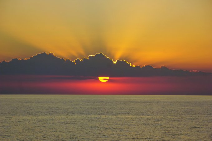 A vibrant sunset over a calm ocean, with the sun partially obscured by a dark cloud layer, casting golden rays across a sky that transitions from yellow to deep pink.