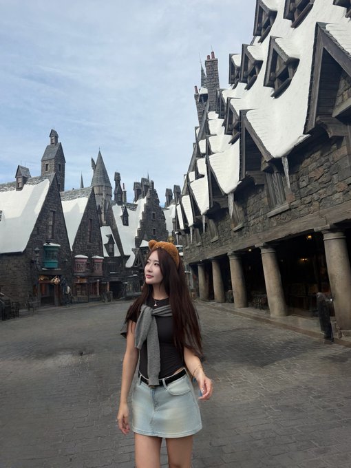 A young woman wearing a brown t-shirt, denim skirt, and an orange headband poses in what appears to be a Harry Potter-themed area with snow-covered medieval-style rooftops.