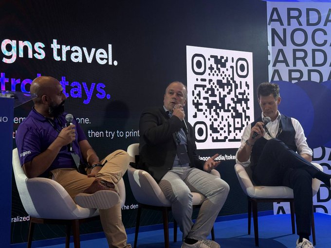 Three men sit on white chairs during a panel discussion; the man in the middle wears a dark jacket while the man on the right holds a microphone, with a large QR code and partial text "igns travel" and "trc...tays" visible on the screen behind them.
