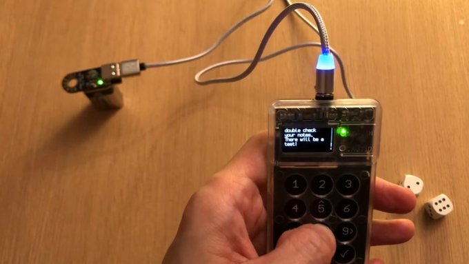 A person holds a transparent DIY handheld device resembling a phone with a numeric keypad and a small screen displaying "double check your roll. you want too a test!", with a USB cable connected to a separate microcontroller and blue LED, while dice sit on the wooden table.