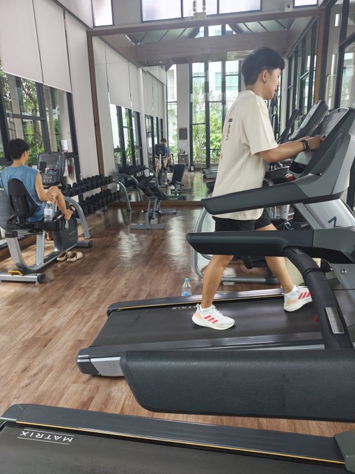 A man in a white t-shirt and shorts exercises on a Matrix treadmill in a gym with wooden floors and large windows, while another person uses exercise equipment in the background.