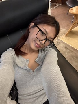A selfie of a young woman wearing black-framed glasses and a light gray button-up henley shirt, sitting on a black leather couch. The post "This is ur onchain opponent" humorously suggests she is a formidable crypto competitor.