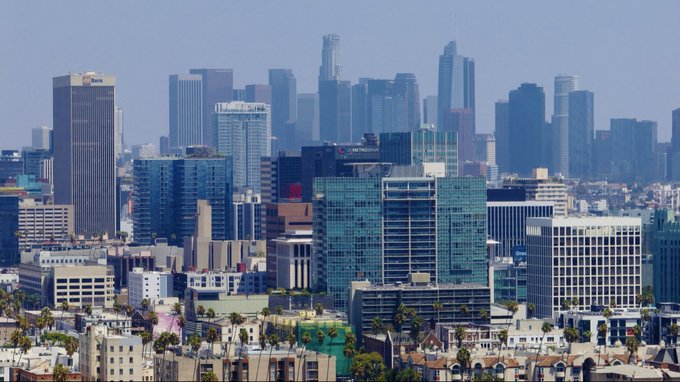 An aerial daytime view of the Los Angeles downtown skyline, showcasing numerous skyscrapers against a hazy sky, with residential buildings and palm trees in the foreground.