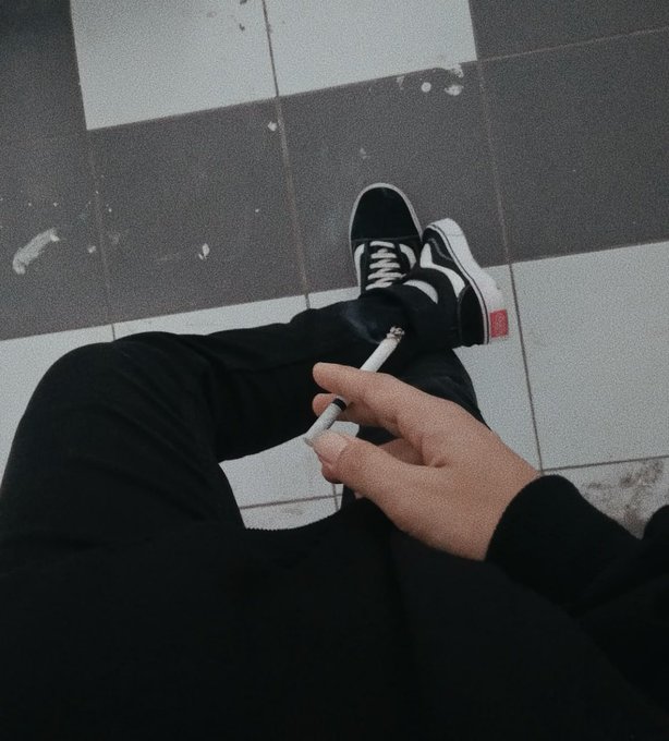 A stylized first-person view of someone sitting down, wearing black pants and black-and-white Vans sneakers, holding a cigarette between their fingers on a gray checkered floor.
