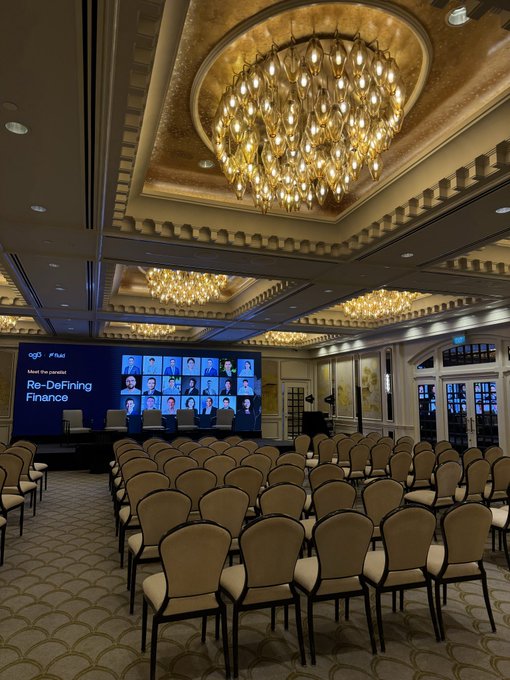 An opulent conference ballroom features ornate gold ceilings, large crystal chandeliers, and rows of empty chairs facing a screen displaying "Re-Defining Finance" with QFC Wealth branding and video participants.