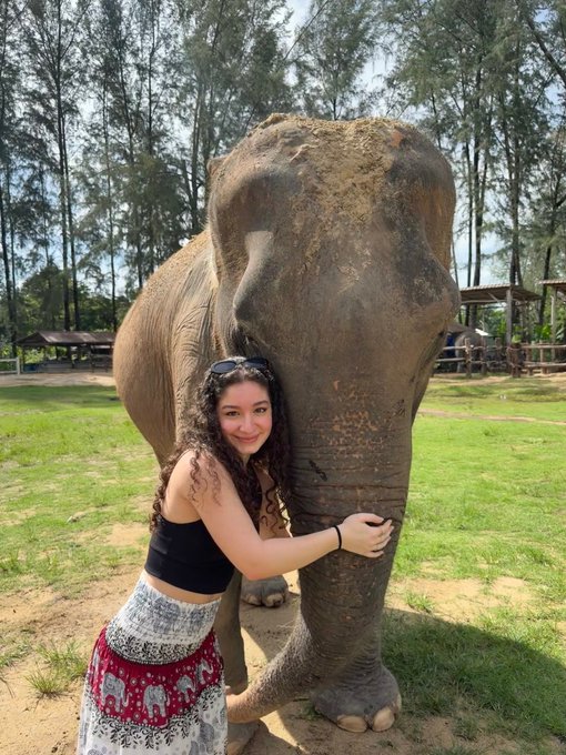 A woman wearing a black tank top and skirt stands next to an elephant, hugging its trunk affectionately as they both stand in front of trees.