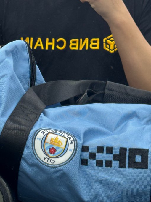 A light blue duffel bag features the Manchester City FC crest and "OKX" sponsor logo, alongside what appears to be a Binance ("BNB CHAIN") item in the background. The image relates to the post's caption about fitness day 20, showcasing crypto exchange merchandise including a backpack, shaker, and towel from Gate.io and OKX/欧意, blending the user's crypto trading lifestyle with their gym routine.