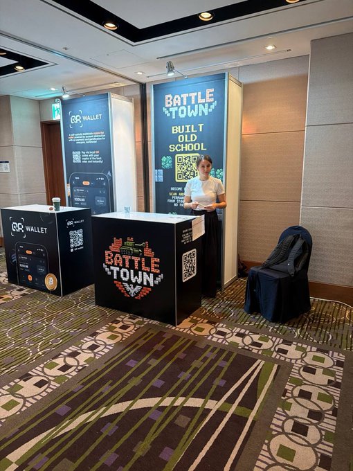 An indoor exhibition booth promotes 'BATTLE TOWN' built in old school style alongside a '68 WALLET' display, featuring pixel-art heart designs, QR codes, and a staff member in a white shirt.