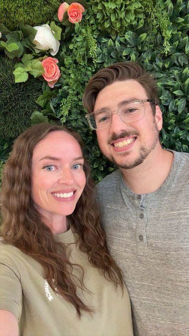 A smiling couple poses closely together in front of a lush green floral wall with pink and white roses. The birthday tribute to “@mikesnft” suggests the man in the photo is being celebrated as the sender’s biggest win in Web3.