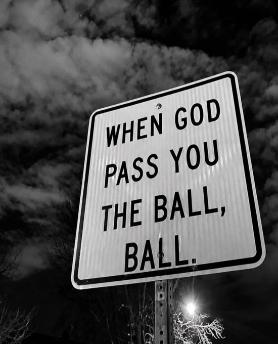A black and white photo of a road sign reading 'WHEN GOD PASS YOU THE BALL, BALL.' against a cloudy sky.