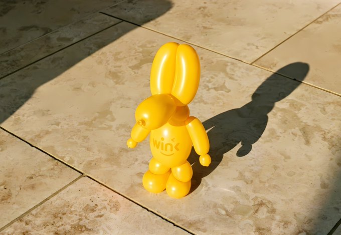 A yellow balloon animal shaped like a bunny with the word 'wink' on its chest, sitting on a tiled floor and casting a shadow in sunlight.