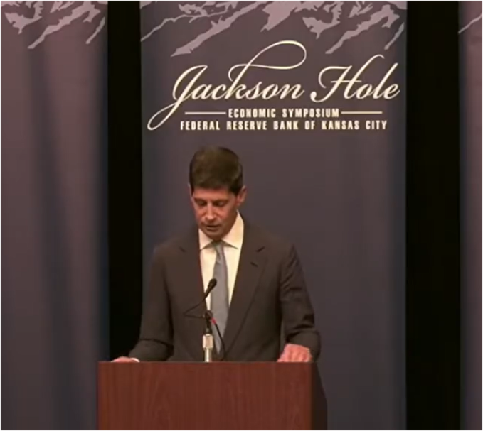 A man in a dark suit and gray tie stands at a podium reading his remarks at the Jackson Hole Economic Symposium, hosted by the Federal Reserve Bank of Kansas City, as shown on the decorative backdrop behind him.
