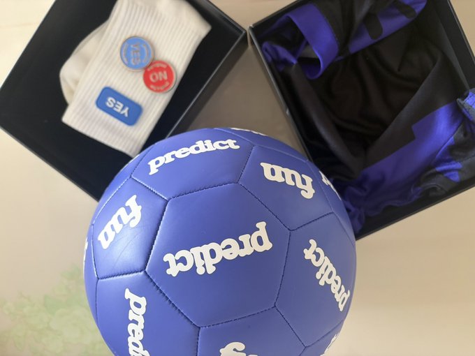 A vibrant purple "predict" branded soccer ball sits in the foreground, accompanied by a gift box containing white socks with "YES" and "ON" patches and a purple and black garment, unboxing the stylish merchandise received from @predictdotfun.