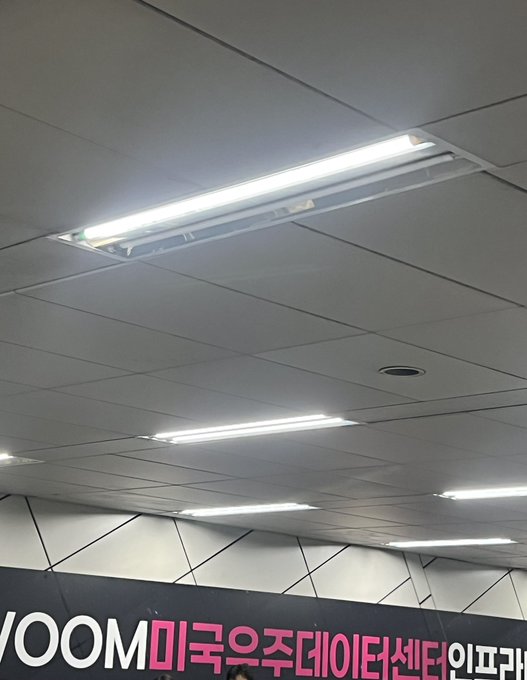 A view of a drop-ceiling with fluorescent lighting and a sign reading "VOOM 미국주데이터센터 인프라" (VOOM US Stock Data Center Infrastructure), suggesting the photo was taken at a financial or data center facility where investment commentary about selling Desenju at its peak was posted.