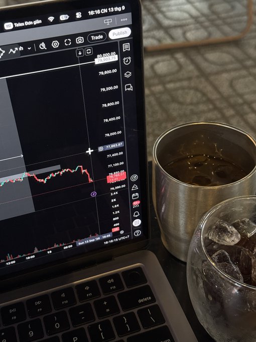 A laptop screen displaying a TradingView bitcoin chart with the price showing around 77,663.71 on the BTCUSD pair, placed next to a metal cup with a dark beverage and a glass with ice.