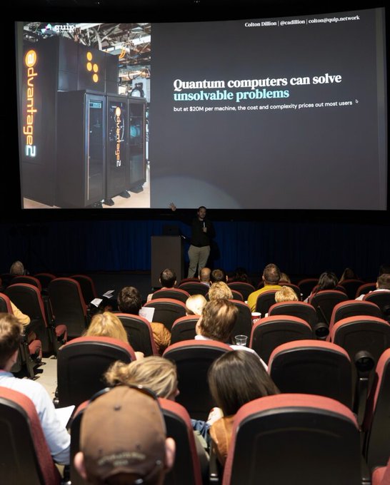 A presenter stands on stage pointing toward a large screen displaying an image of a D-Wave Advantage 2 quantum computer, with text stating "Quantum computers can solve unsolvable problems - but at $20M per machine, the cost and complexity prices out most users." The presentation appears to be giving context for the QUIP network ($QUIP) project, explaining why decentralized quantum computing matters as a solution to the prohibitive cost and accessibility barriers of traditional quantum machines.