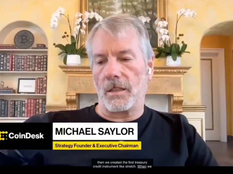 The image features a man wearing a black shirt, sitting in front of a fireplace with potted plants around him. The man appears to be talking about cryptocurrency or investing in Bitcoin. The text "Michael Saylor" can also be seen on his shirt.