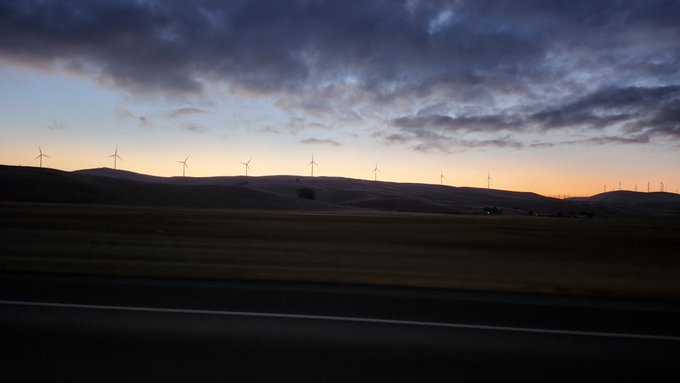Silhouettes of wind turbines along rolling hills against a cloudy sunset sky, viewed from a road.