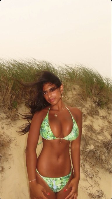 A woman with long dark hair poses in a green floral bikini on a sandy beach with dune grass behind her.