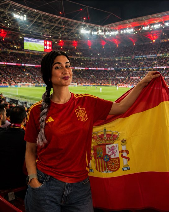 A woman in a red Spain national football team jersey holds a Spanish flag at a crowded stadium during a match.