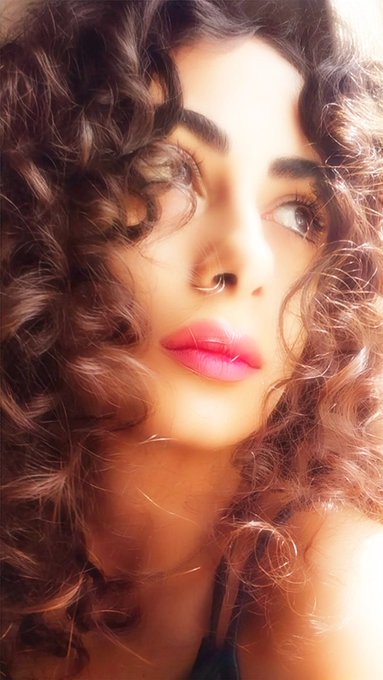 A close-up portrait of a young woman with voluminous curly brown hair, pink lipstick, and a nose ring, looking off to the side.