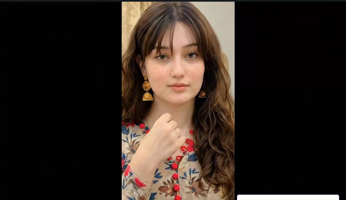 The image features a woman with long brown hair wearing a floral shirt, posing for a picture. She has gold earrings that catch the light, adding an elegant touch to her appearance. The background of the photo is black, which contrasts nicely with the woman's colorful attire and highlights her as the main subject in the image.