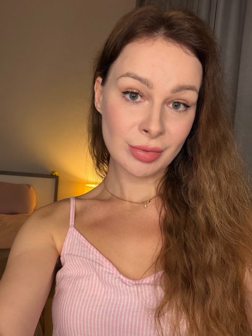 A selfie of a young woman with long auburn hair, wearing a pink ribbed camisole and a delicate gold necklace, in a softly lit bedroom setting.