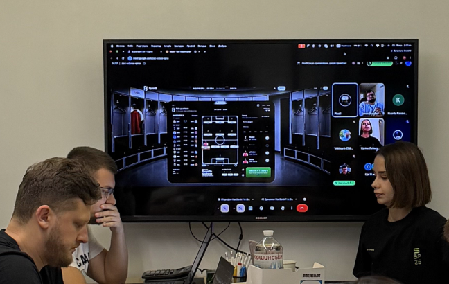 Three people are gathered around a large wall-mounted monitor displaying a virtual event or livestream interface with multiple participant thumbnails visible.