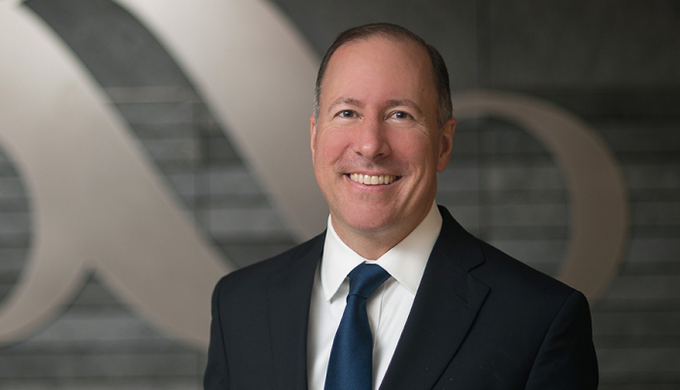 A professional headshot of a smiling man in a dark suit and blue tie, standing in front of a blurred gray background with a large '60' logo.