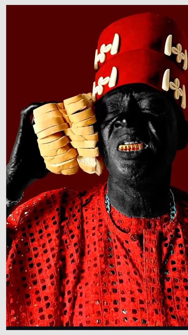 A stylized portrait of an older man with black skin wearing a vibrant red traditional outfit and a tall red hat with bone-like decorations. He holds a bunch of yellow bananas near his face, which features gold teeth, against a deep red background.