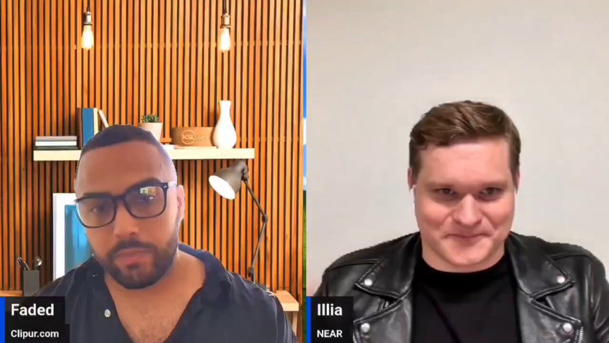 The image shows two men sitting next to each other with their faces displayed on a computer screen during an interview or discussion. The man on the left has his face partially visible, while the man on the right has his face fully visible. They are both wearing glasses and have short hair. The background of the image is white, which contrasts with the brown wooden wall behind them. There are also several books scattered around the room, indicating a casual or informal setting for their conversation.