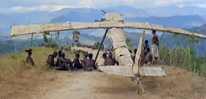 A group of people gather on a grassy hilltop around a primitive handmade glider/aircraft with fabric-covered wings, set against a backdrop of distant blue mountains.
