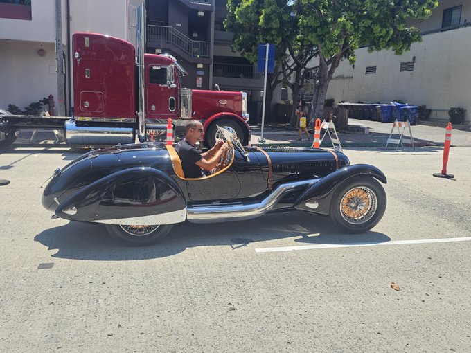 A black classic convertible sports car parked on a street with a man seated inside, while a maroon semi-truck and orange traffic cones are visible in the background.