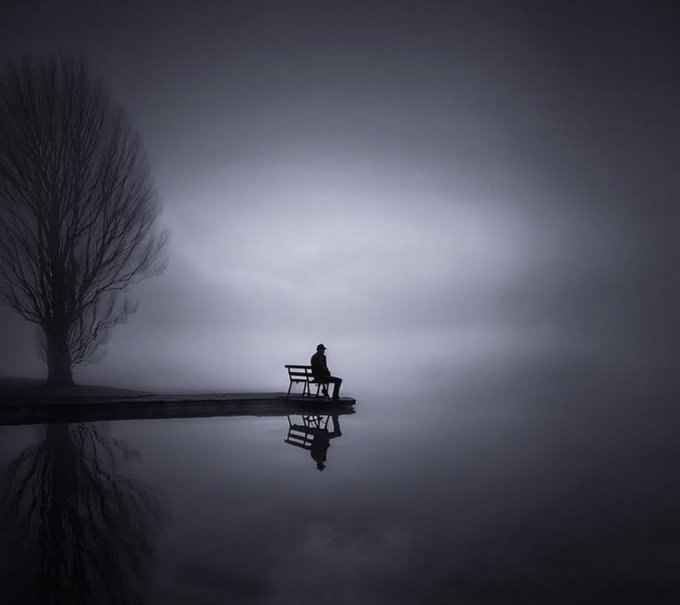 A moody, atmospheric photograph of a solitary figure sitting on a bench by a misty lake, with a bare tree on the left and the scene's reflection visible in the calm water.